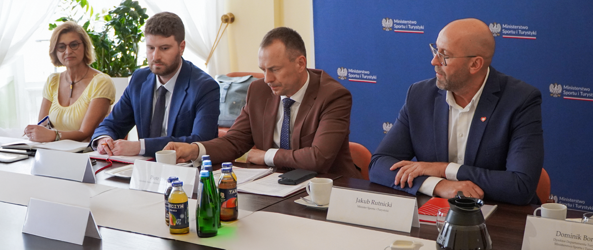 Minister Jakub Rutnicki oraz Sekretarz Stanu Piotr Borys podczas spotkania z branżą turystyczną, siedzą za stołem na tle niebieskiej ścianki z logiem ministerstwa